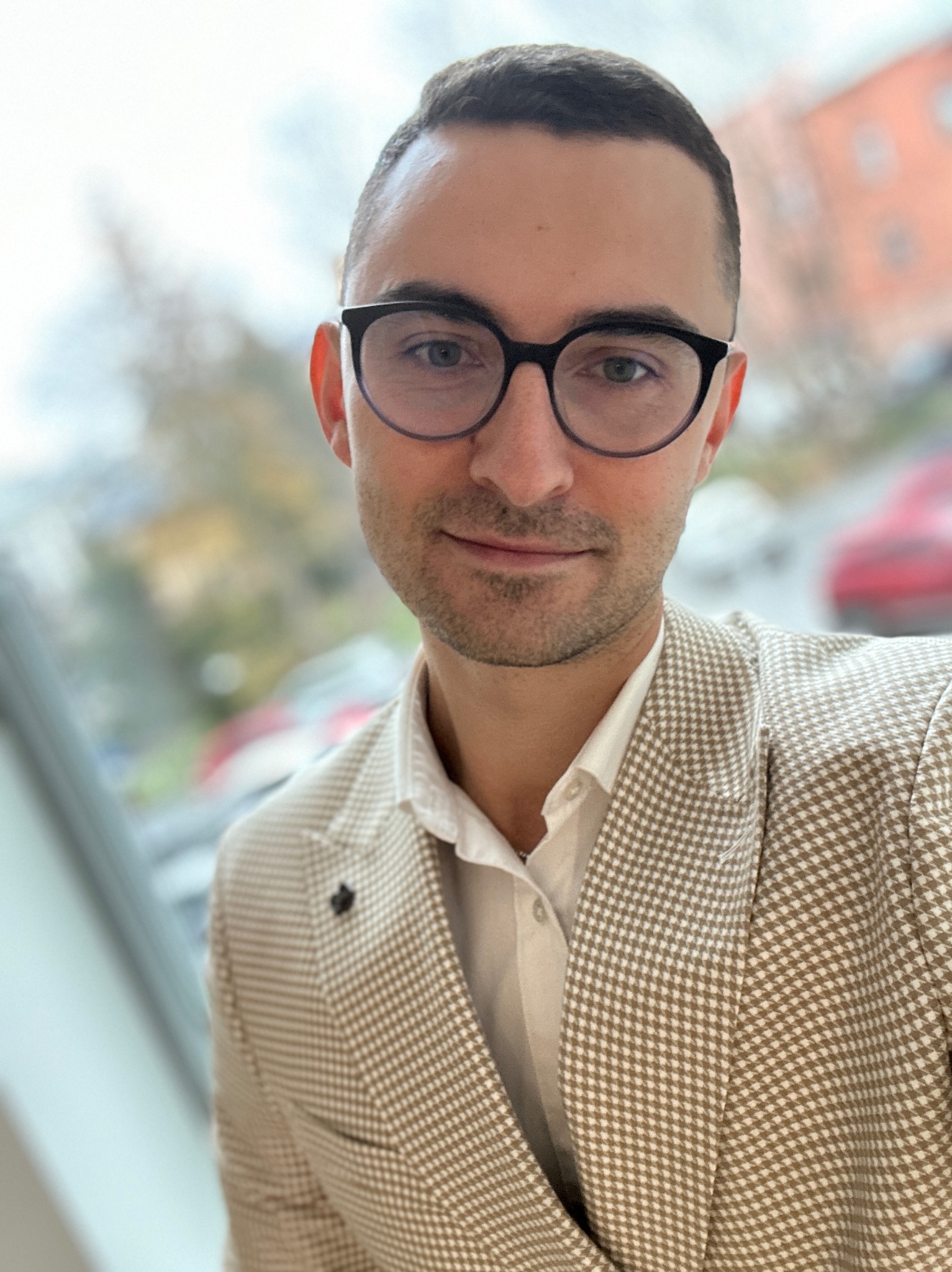 Portret mężczyzny w okularach i beżowej marynarce w drobną kratę, stojącego na tle okna z widokiem na budynek i samochody.