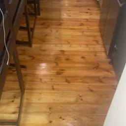 Świeżo polakierowana, błyszcząca drewniana podłoga z widocznymi słojami i sękami, częściowo zasłonięta przez meble kuchenne i krzesła barowe.