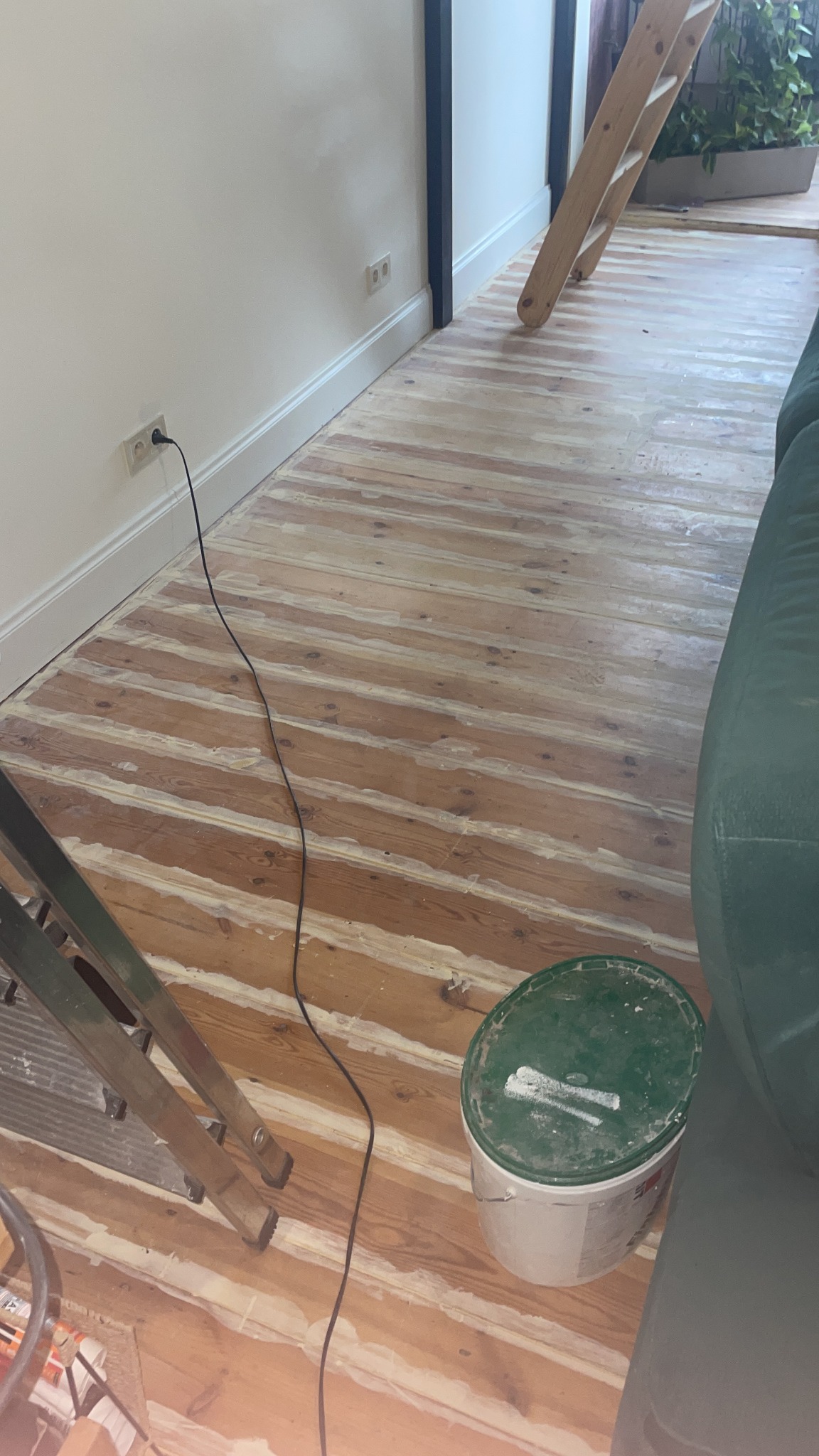 Widok z góry na drewnianą podłogę w trakcie renowacji, z widocznymi szczelinami wypełnionymi świeżą masą szpachlową, drabina malarska i wiadro z farbą w tle, kabel zasilający podłączony do gniazdka...