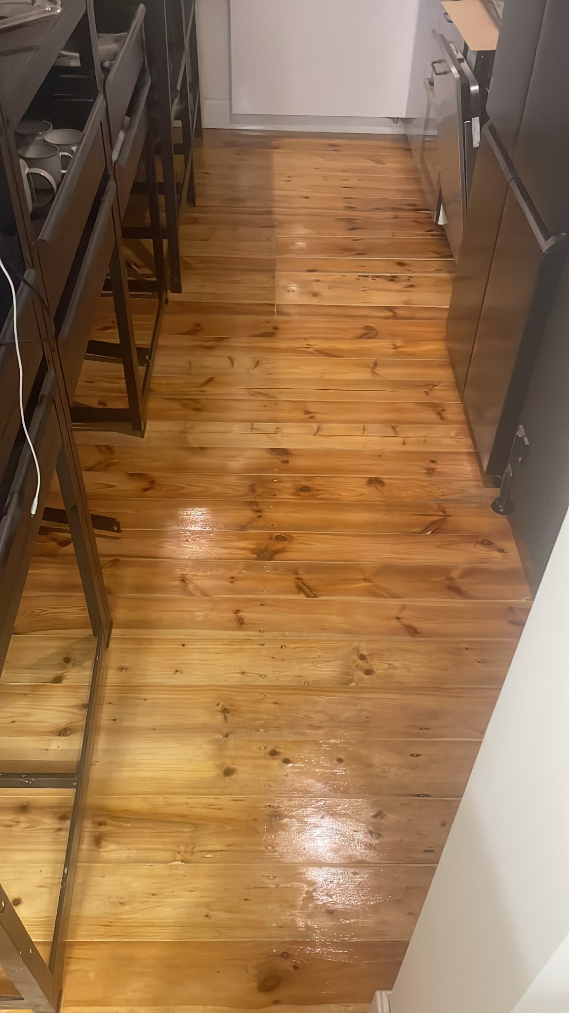 Świeżo polakierowana, błyszcząca drewniana podłoga z widocznymi słojami i sękami, częściowo zasłonięta przez meble kuchenne i krzesła barowe.