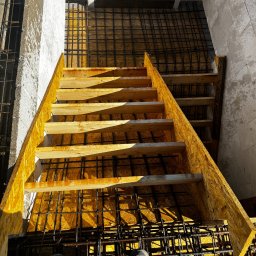 ELITE ENGINEERING SPÓŁKA Z OGRANICZONĄ ODPOWIEDZIALNOŚCIĄ - Szczegółowe ujęcie z góry na szalunek schodów zbrojonych, przygotowany do zalania betonem, widoczne buty osoby stojącej na zbrojeniu.