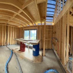 ELITE ENGINEERING SPÓŁKA Z OGRANICZONĄ ODPOWIEDZIALNOŚCIĄ - Wnętrze domu w trakcie budowy: drewniana konstrukcja szkieletowa, instalacje w podłodze, okna z folią ochronną, widoczne elementy instalacji elektrycznej i hydraulicznej.