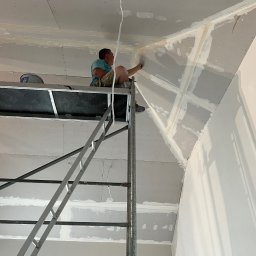ELITE ENGINEERING SPÓŁKA Z OGRANICZONĄ ODPOWIEDZIALNOŚCIĄ - Pracownik na rusztowaniu szpachluje łączenia płyt gipsowo-kartonowych na skośnym suficie poddasza, wykończenie wnętrza.