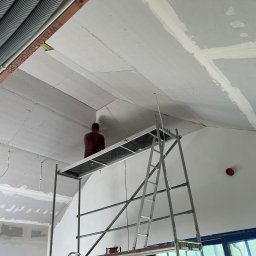 ELITE ENGINEERING SPÓŁKA Z OGRANICZONĄ ODPOWIEDZIALNOŚCIĄ - Praca na wysokości: mężczyzna na rusztowaniu montuje płyty gipsowo-kartonowe na skośnym suficie poddasza, widoczne łączenia płyt i rury instalacyjne.