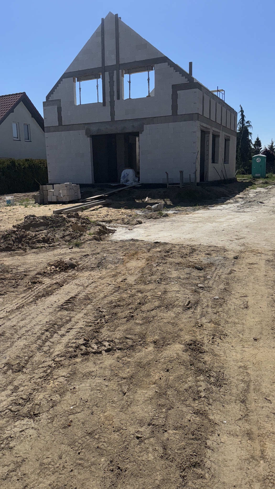 Surowy stan domu z betonu komórkowego w trakcie budowy. Widoczne otwory okienne, drzwiowe i teren budowy z ziemią oraz materiałami budowlanymi.