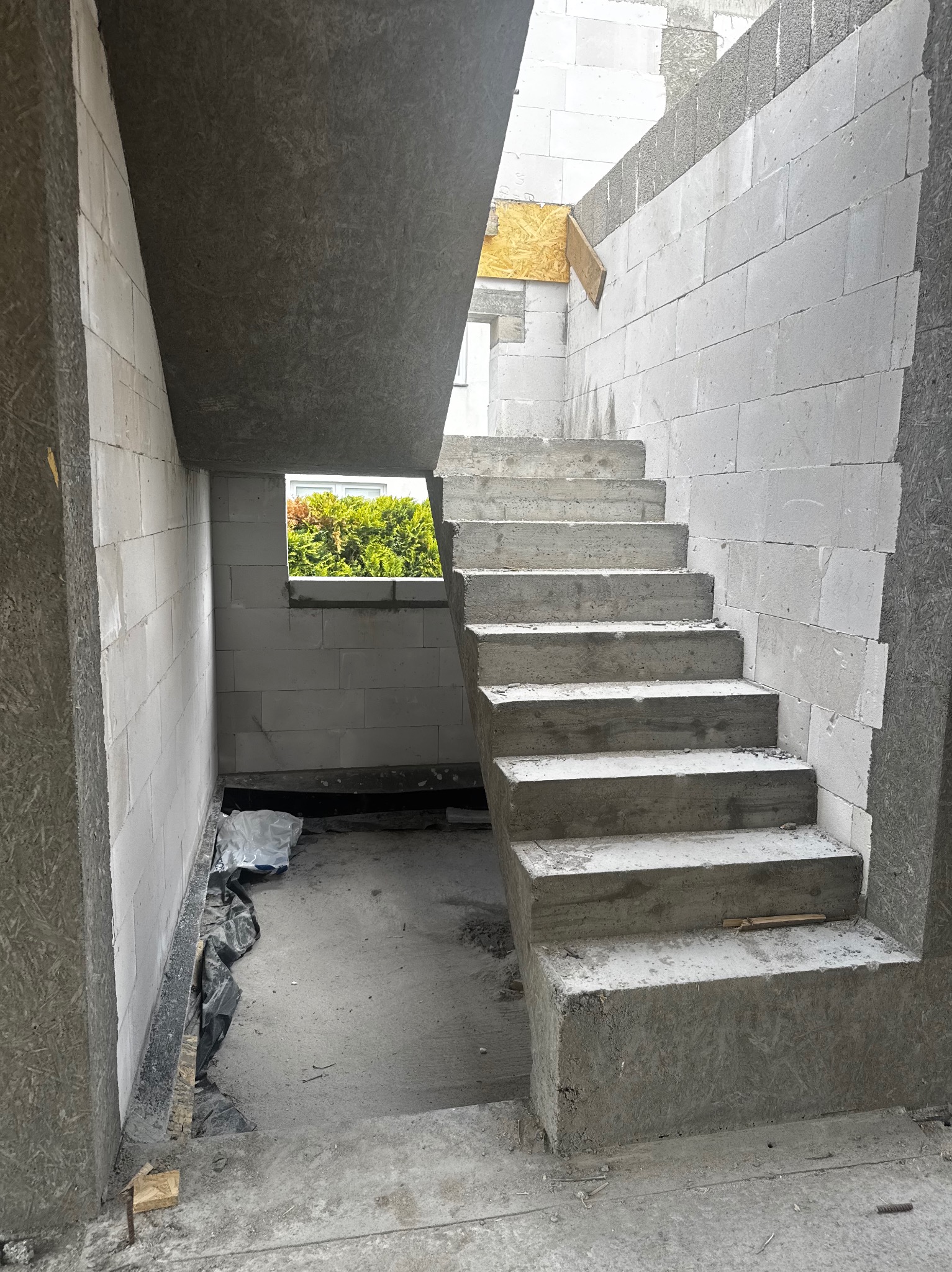 Surowe betonowe schody wewnątrz budynku w trakcie budowy, widok na okno z zielenią, ściany z białych bloczków, szalunki.