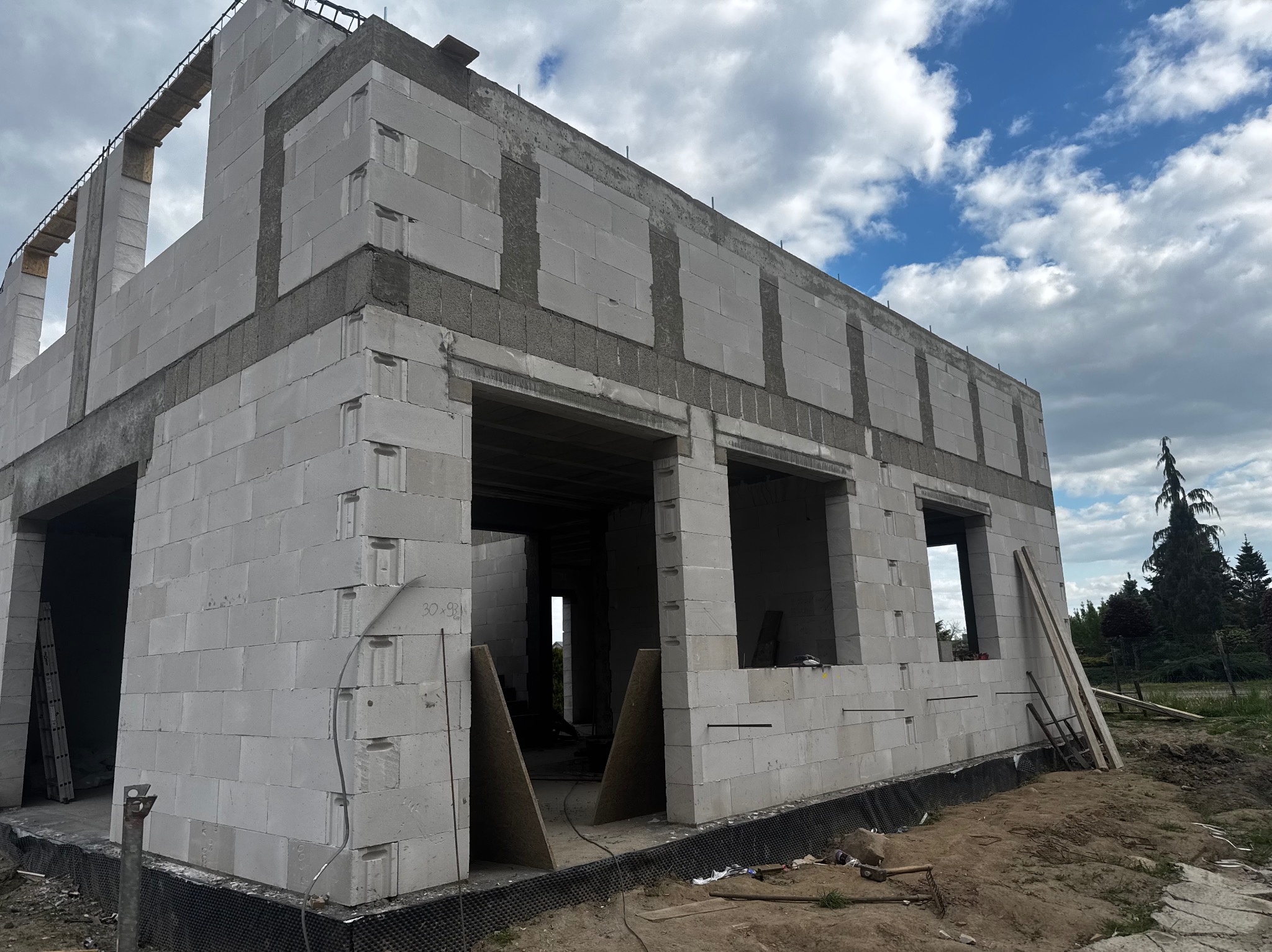 Surowy stan domu z betonu komórkowego, widoczne otwory okienne i drzwiowe, zbrojenia, teren budowy. Budynek w trakcie wznoszenia, widok z dołu na tle pochmurnego nieba.