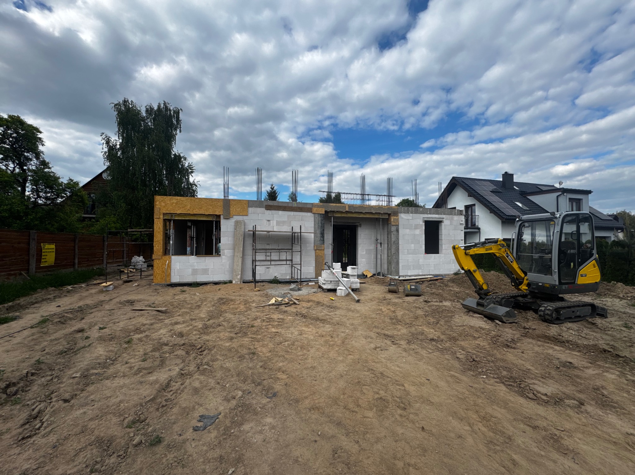 Budowa domu jednorodzinnego w stanie surowym otwartym, widoczny mini-koparka na tle nieba z chmurami i sąsiednich budynków.