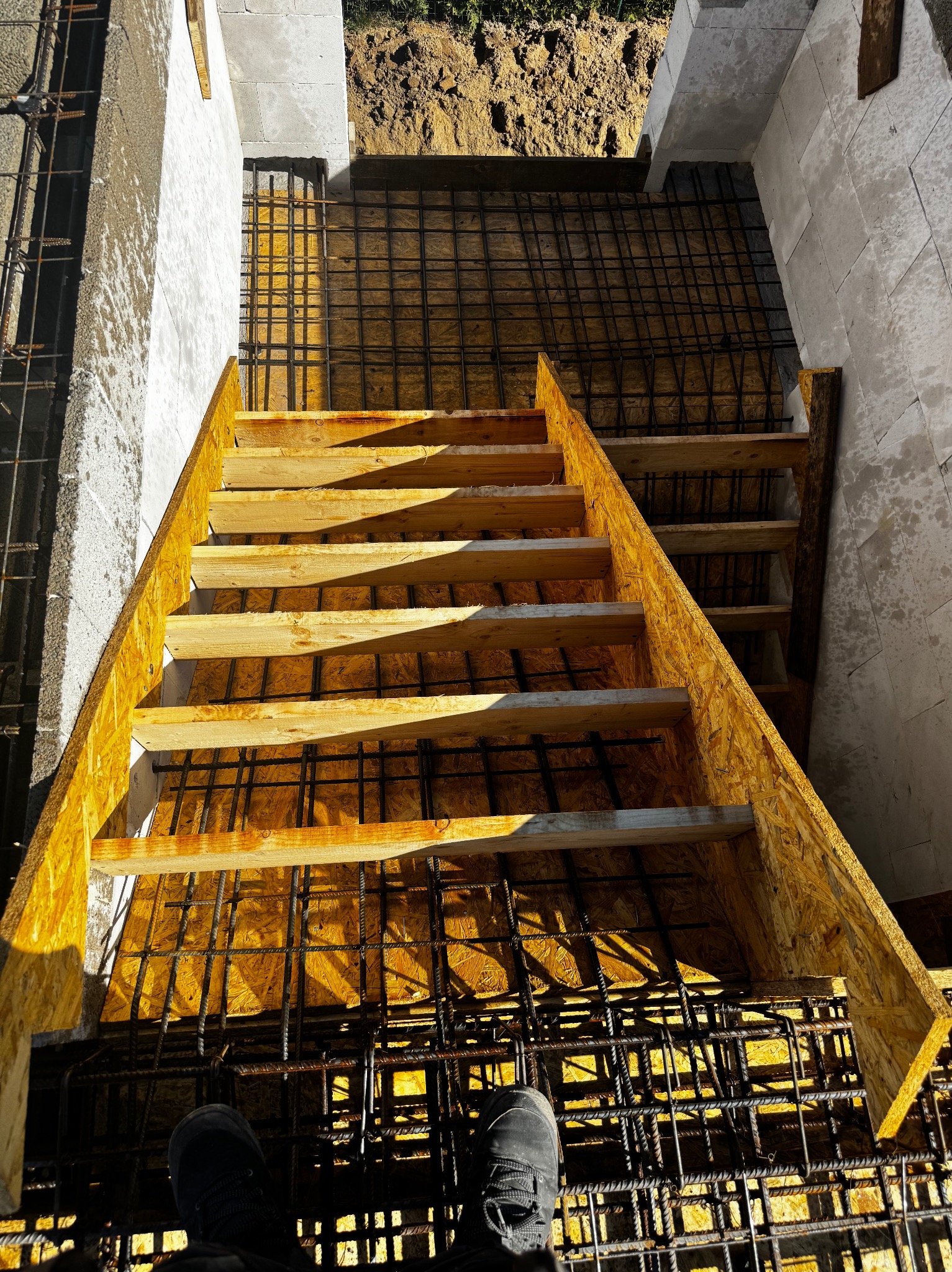 Szczegółowe ujęcie z góry na szalunek schodów zbrojonych, przygotowany do zalania betonem, widoczne buty osoby stojącej na zbrojeniu.