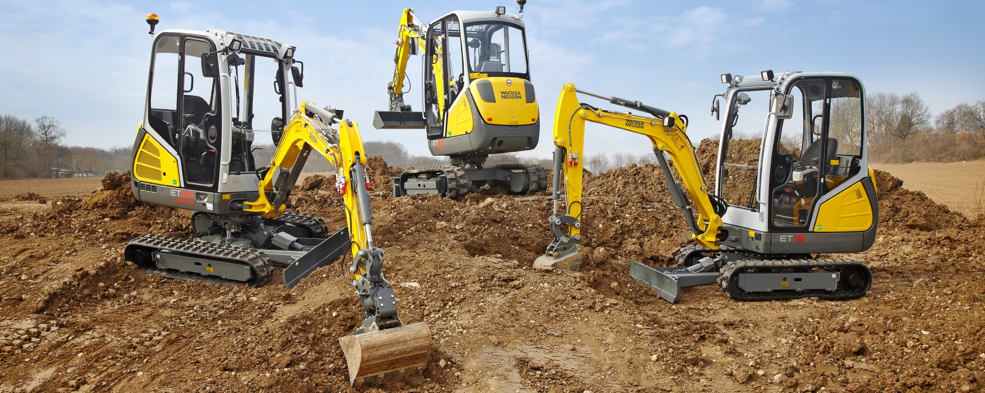 Trzy żółto-szare mini koparki Wacker Neuson na nierównym terenie. Koparki w różnych pozycjach, z widocznymi łyżkami i gąsienicami, na tle błękitnego nieba.