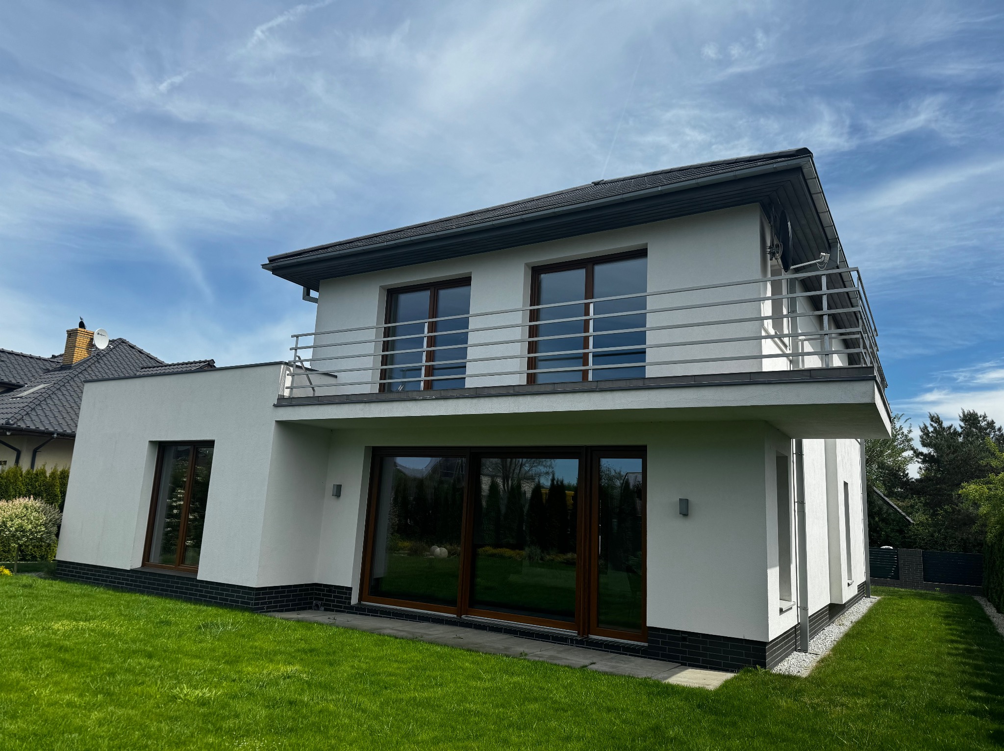 Dwupiętrowy, biały dom z balkonem i dużymi oknami, otoczony zielonym trawnikiem pod błękitnym niebem. Widoczne detale architektoniczne i staranne wykończenie.