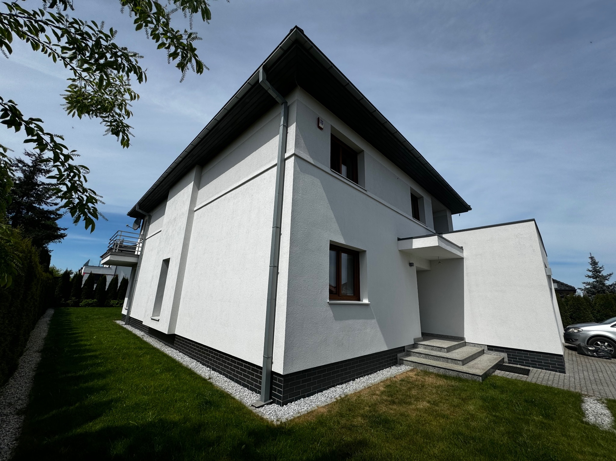 Biały, dwukondygnacyjny dom z ciemną podmurówką i szarymi schodami wejściowymi, widziany z perspektywy ogrodu w słoneczny dzień. Minimalistyczna architektura.