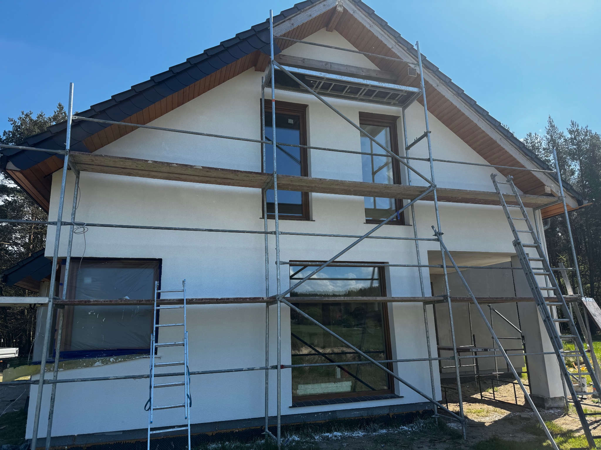Biały dom z ciemnobrązowymi oknami i ciemnym dachem w trakcie wykończenia, otoczony rusztowaniem. Jeden z okien jest zakryty folią.