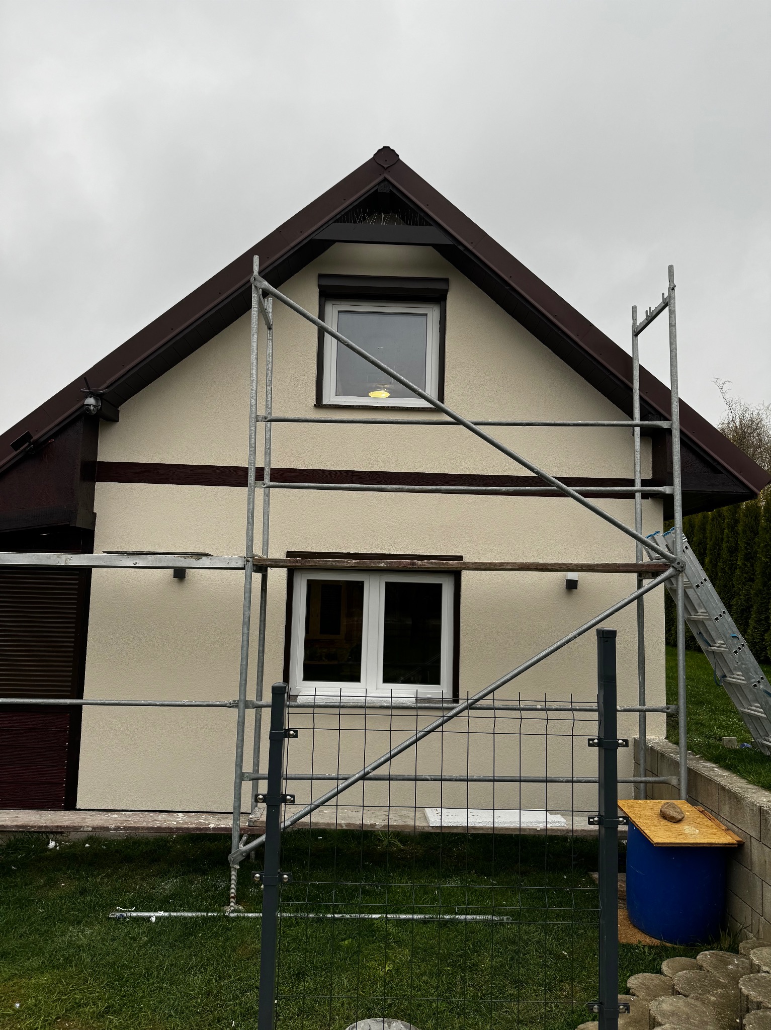 Dom jednorodzinny z beżową elewacją, brązowym dachem i ramami okiennymi, częściowo zasłonięty rusztowaniem. Widoczna drabina oparta o ścianę budynku.
