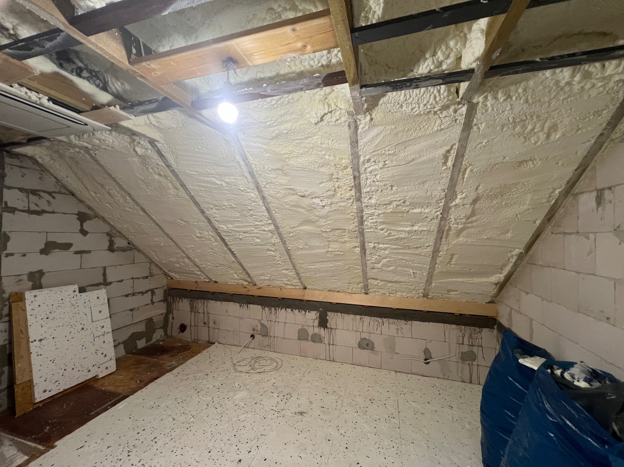 Ocieplenie poddasza pianą PUR, widoczne krokwie i bloczki betonowe. Izolacja natryskowa na skośnym dachu, biała piana wypełnia przestrzeń między elementami konstrukcyjnymi.