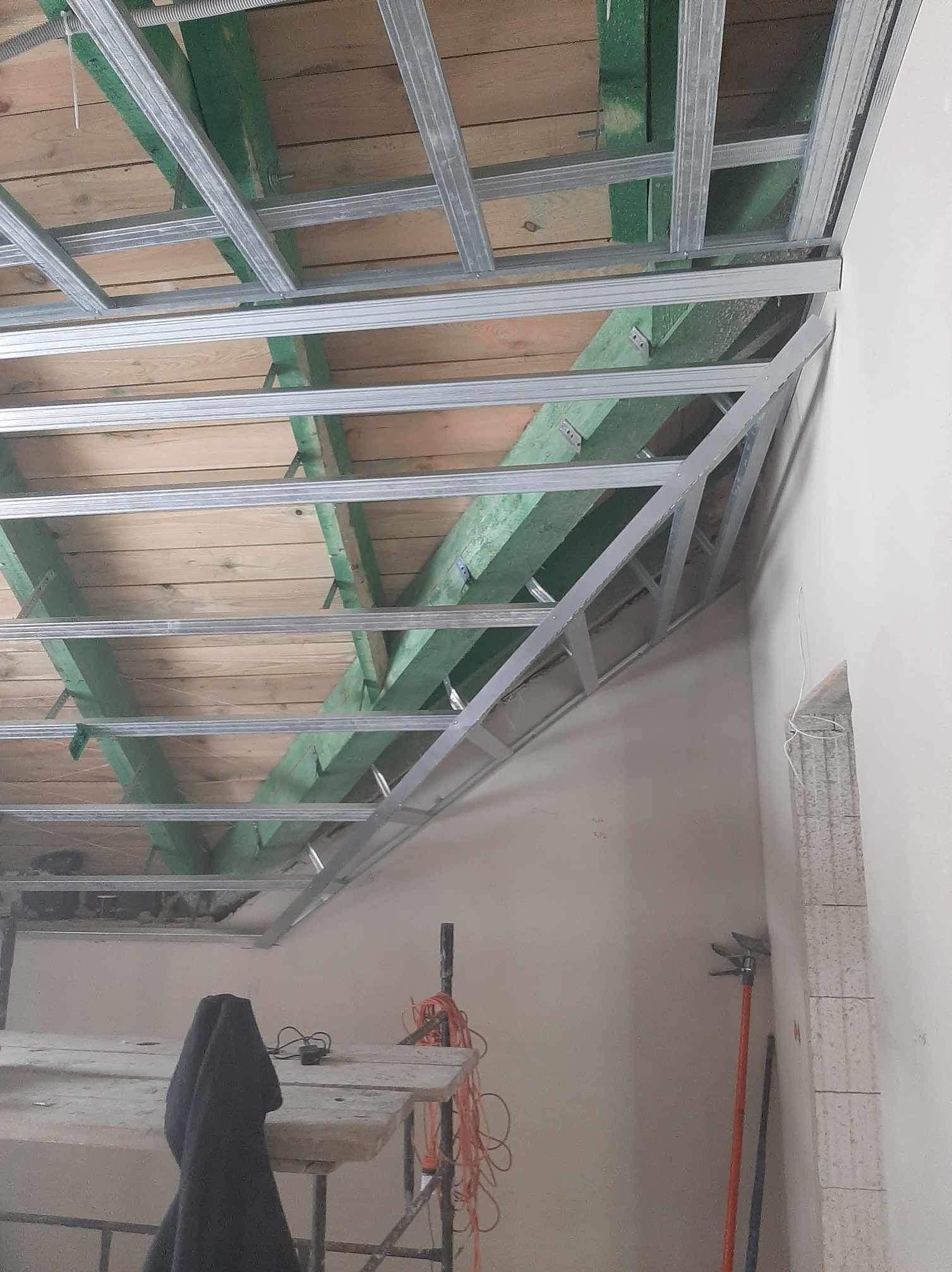 Konstrukcja sufitu podwieszanego z profili metalowych, widok z dołu na drewnianą konstrukcję dachu i część ściany. Widoczny fragment rusztowania.