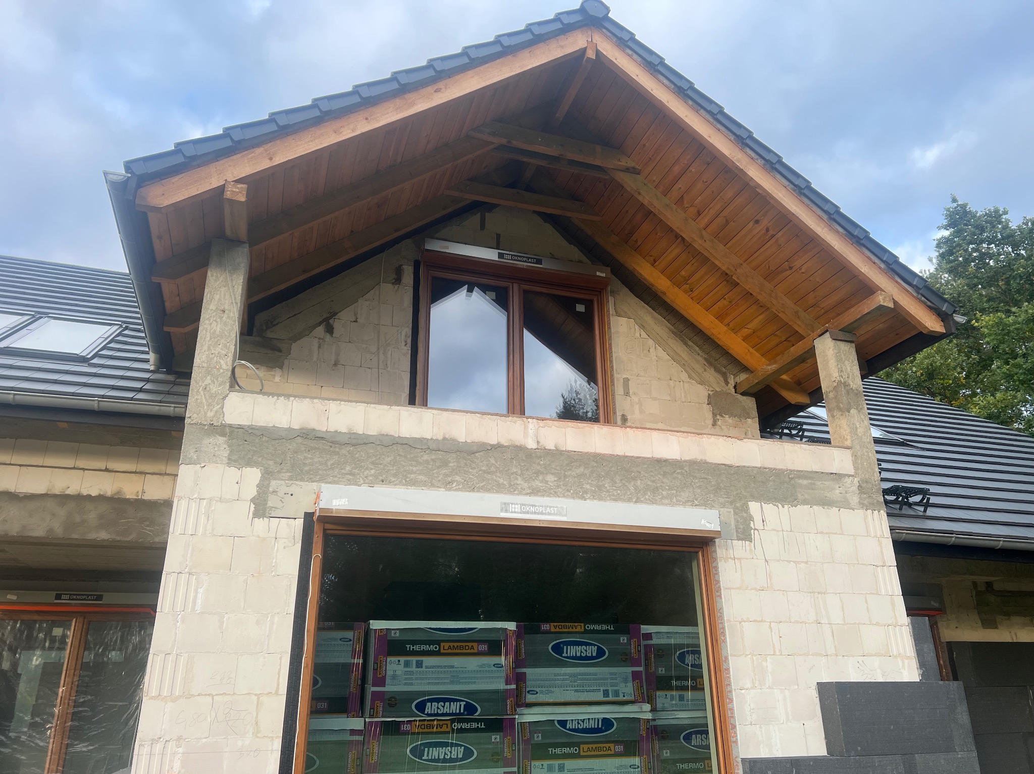 Surowy stan domu z pustaków, widoczne drewniane elementy dachu i okna. Na dole palety z materiałami izolacyjnymi. Budynek w trakcie budowy.