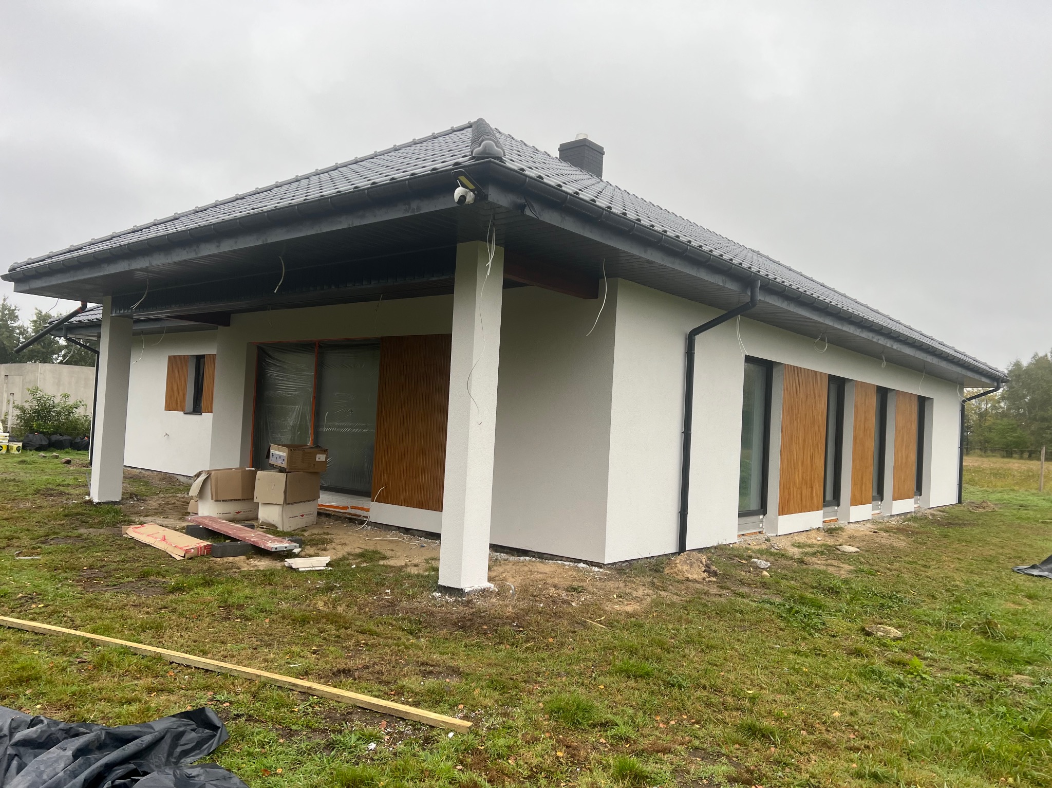 Niedokończony parterowy dom z białym tynkiem i grafitową dachówką. Widoczne kartony i materiały budowlane wokół budynku. Okna częściowo zabezpieczone folią.