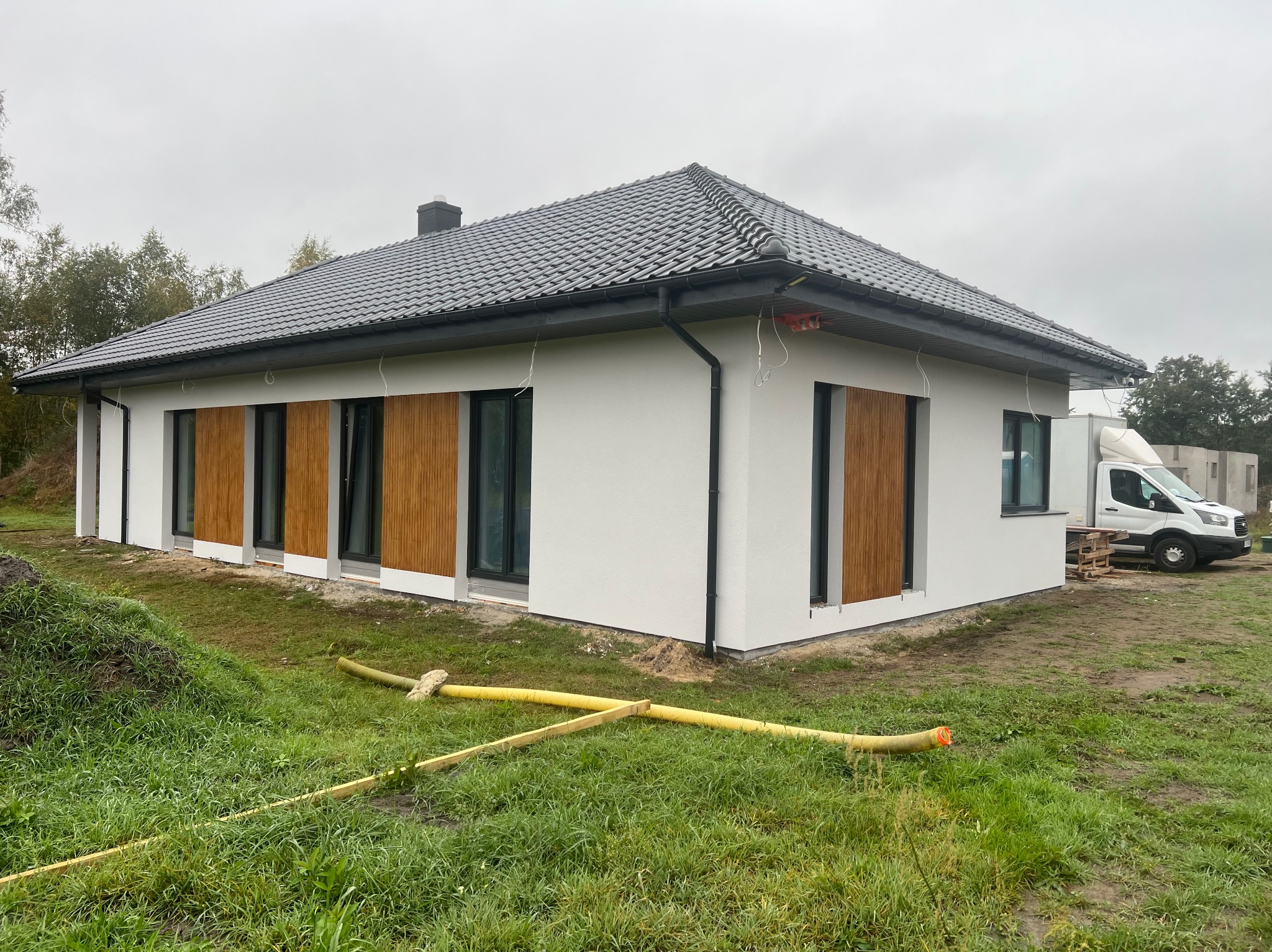 Niedawno wybudowany, parterowy dom z szarą dachówką i drewnianą okładziną. Widoczny biały bus i teren wokół domu w trakcie zagospodarowywania. Pochmurne niebo.