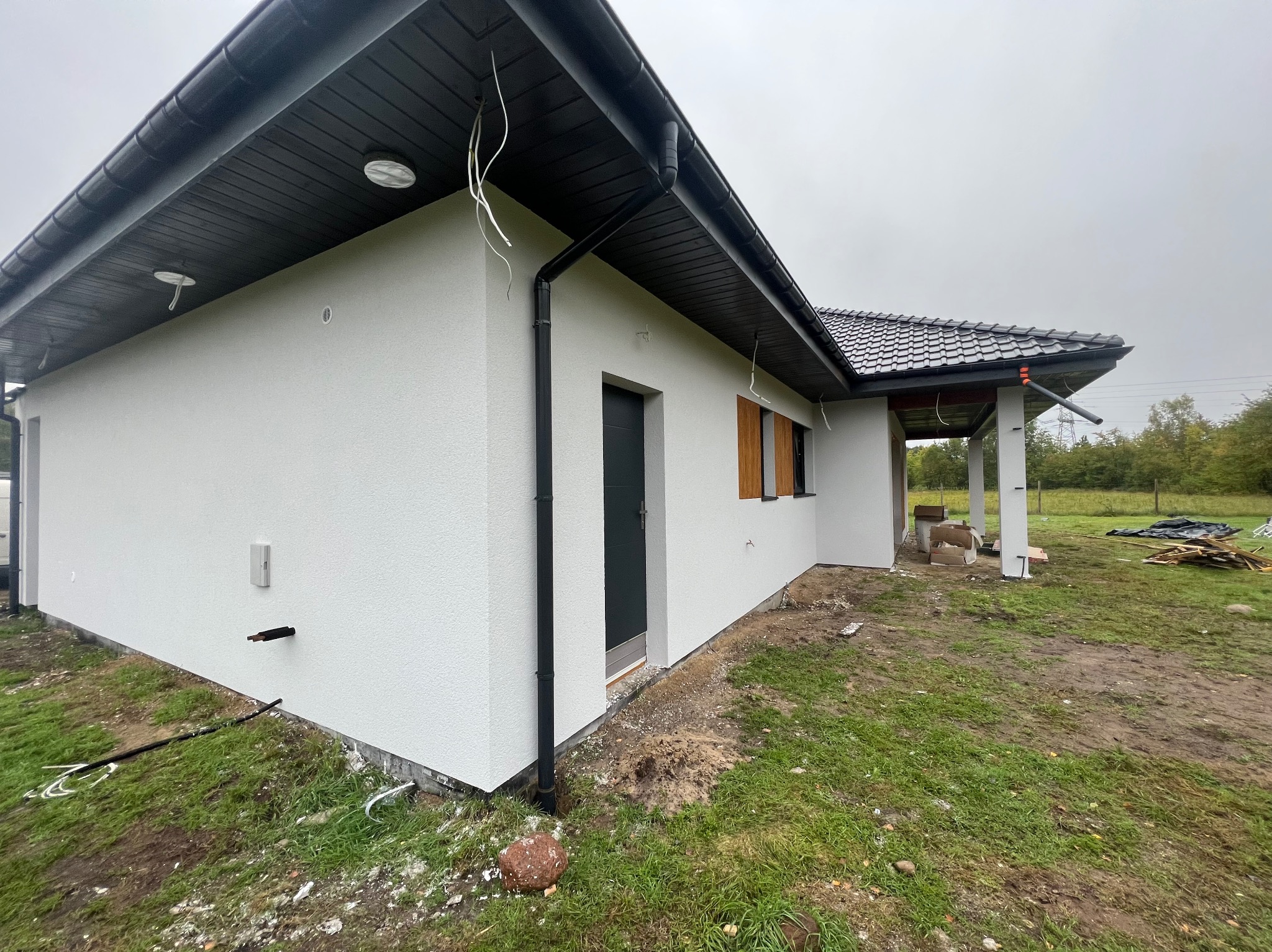 Nowoczesny, parterowy dom z białą elewacją i ciemną dachówką. Widoczne rynny, okna z drewnianymi okiennicami i elementy instalacji na zewnątrz budynku.