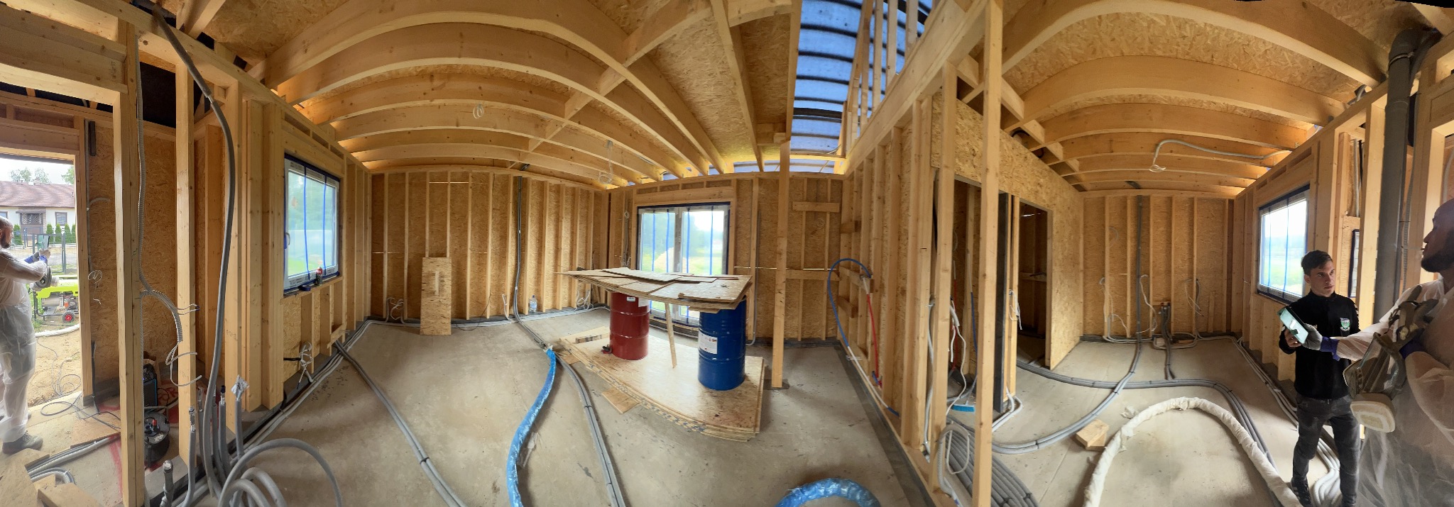 Wnętrze domu w trakcie budowy: drewniana konstrukcja szkieletowa, instalacje w podłodze, okna z folią ochronną, widoczne elementy instalacji elektrycznej i hydraulicznej.