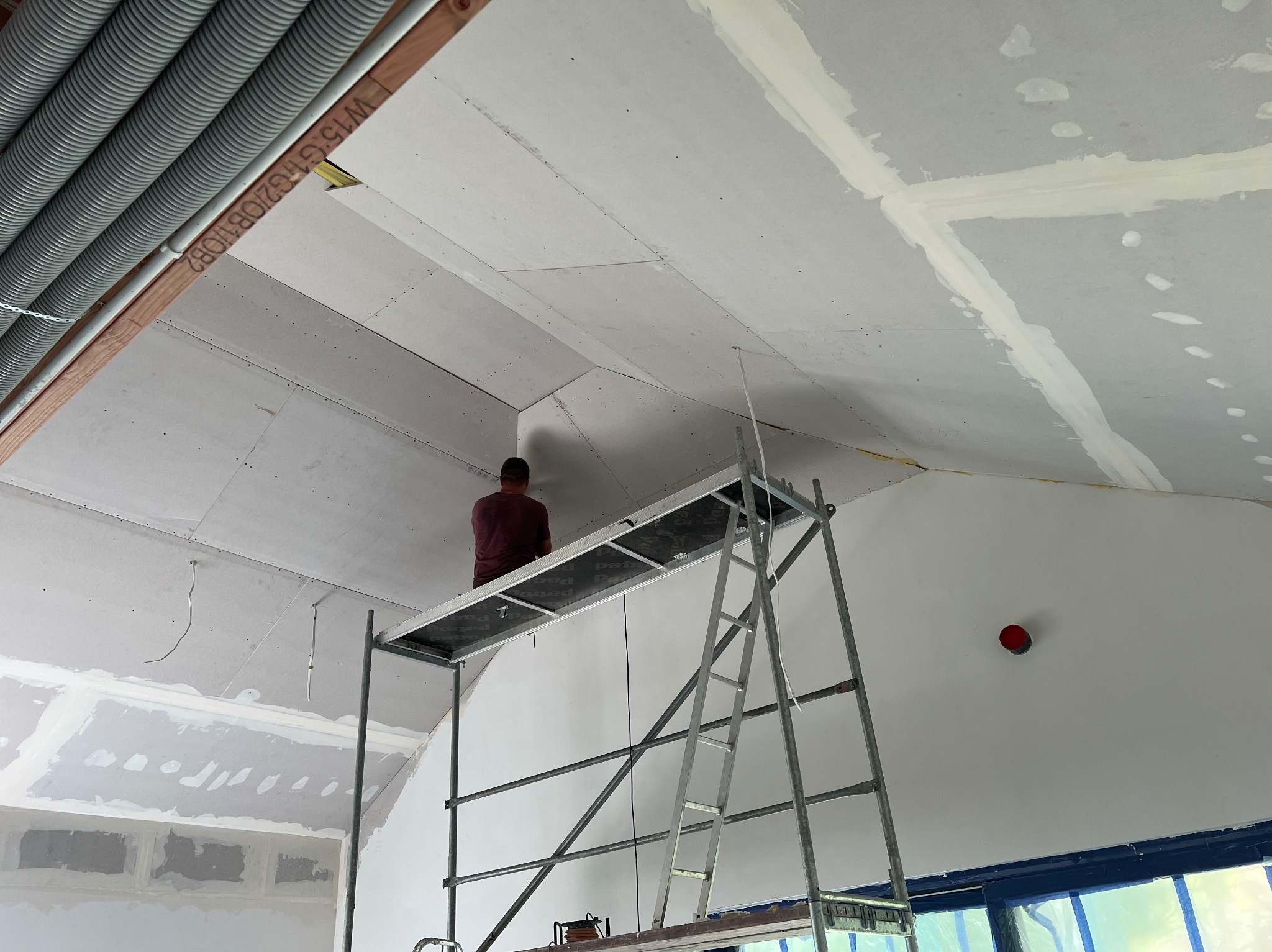 Praca na wysokości: mężczyzna na rusztowaniu montuje płyty gipsowo-kartonowe na skośnym suficie poddasza, widoczne łączenia płyt i rury instalacyjne.