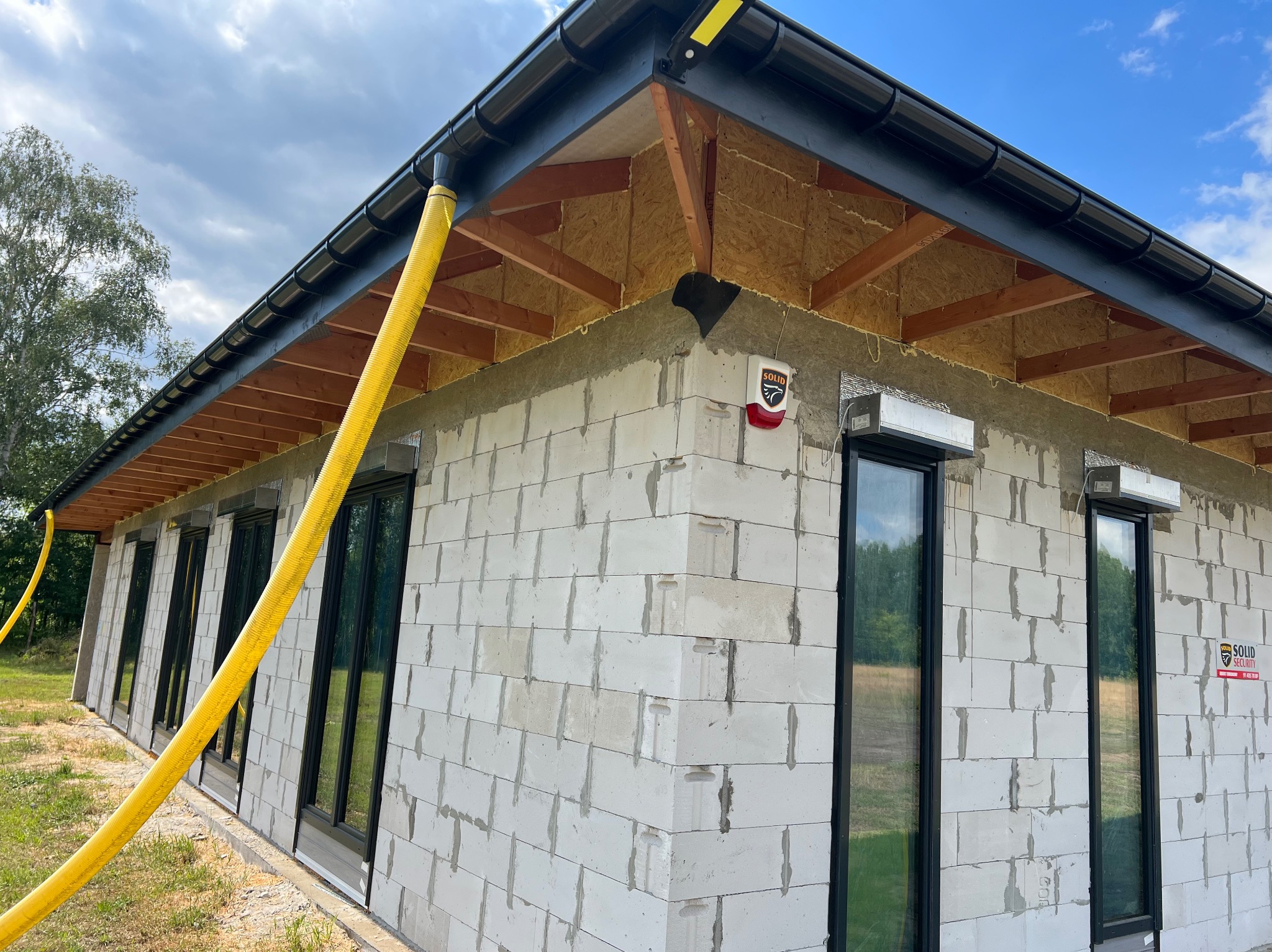 Surowy stan domu z białych bloczków, widoczne okna z czarnymi ramami, orynnowanie i żółty przewód wentylacyjny. Budynek w trakcie budowy, z elementami alarmu.