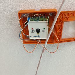 SAPinstal Mariusz Sikora - Pomarańczowa obudowa alarmowa z widocznym modułem sterującym, przewodami i przyciskiem OK, zamontowana na białej, teksturowanej ścianie. Widoczny fragment rurki instalacyjnej.