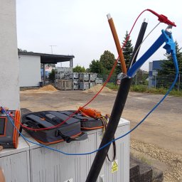 SAPinstal Mariusz Sikora - Pomiary elektryczne: kabel z podłączonymi przewodami pomiarowymi i miernikiem na zewnątrz budynku. Widoczne narzędzia i skrzynki narzędziowe.