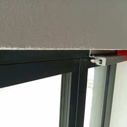 SAPinstal Mariusz Sikora - Zamontowany napęd elektryczny okna, widoczny mechanizm i przewody. Ciemna rama okienna kontrastuje z jasnym tłem. Czerwony element obudowy napędu dodaje akcentu kolorystycznego.