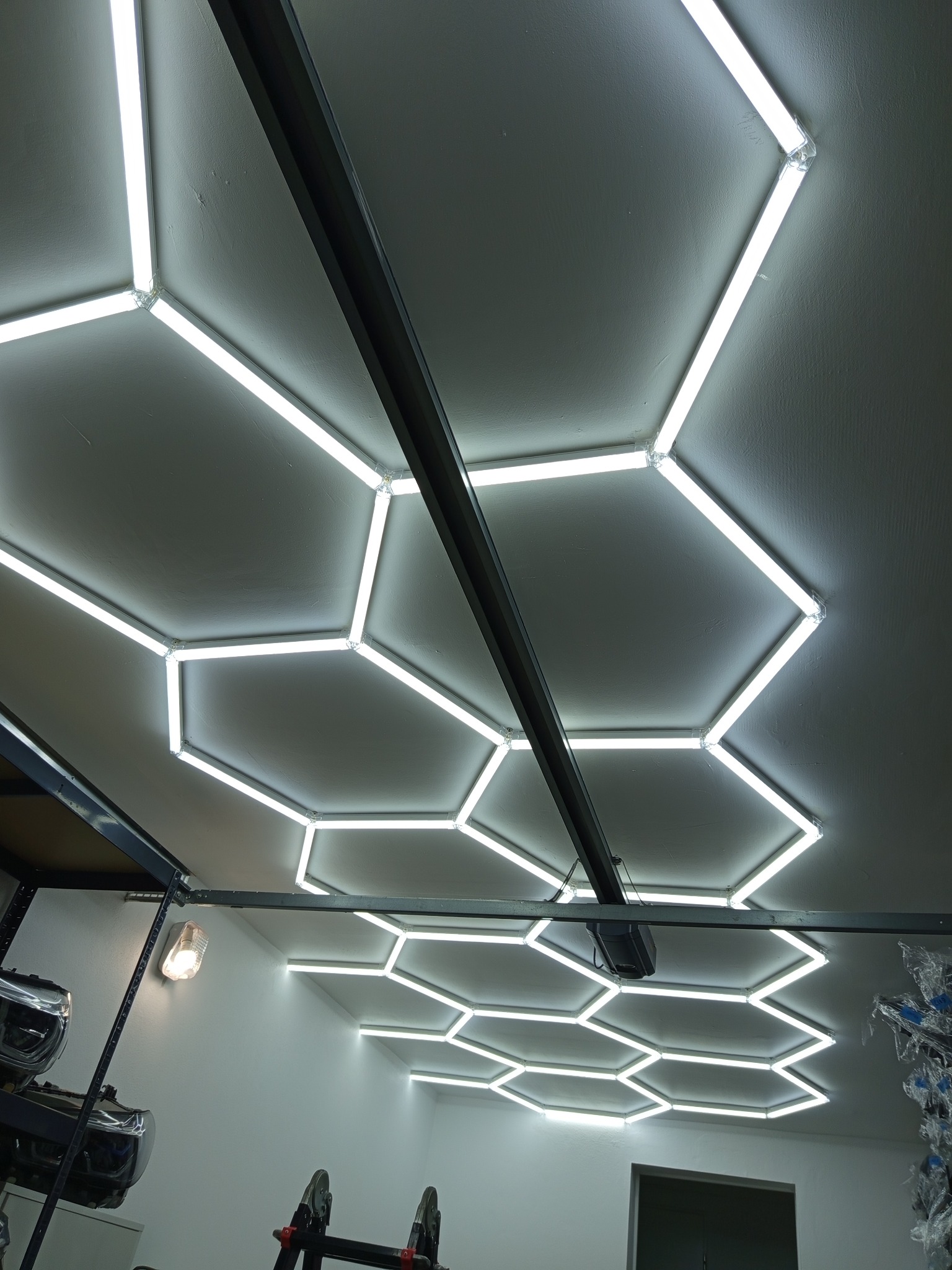 Geometryczna instalacja świetlna LED na suficie garażu, tworząca wzór plastra miodu. Widoczna metalowa konstrukcja i oświetlenie pomocnicze. Nowoczesne oświetlenie.