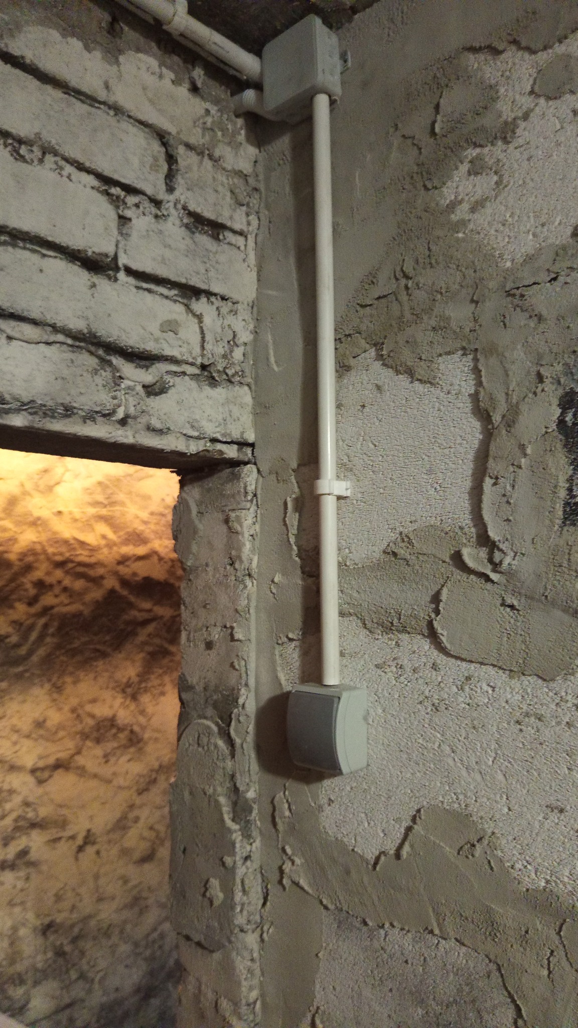 Szara rurka instalacyjna z puszką na ścianie z surowym tynkiem, widoczna część muru z cegły i otwór w ścianie z jasnym wnętrzem. Instalacja w rogu pomieszczenia.