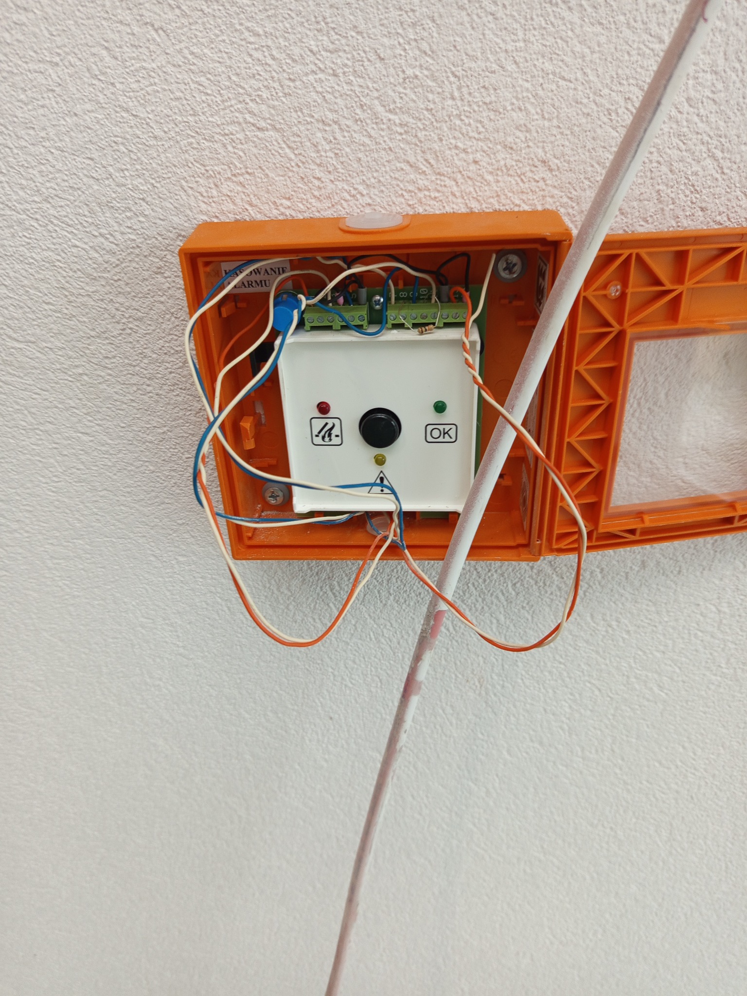 Pomarańczowa obudowa alarmowa z widocznym modułem sterującym, przewodami i przyciskiem OK, zamontowana na białej, teksturowanej ścianie. Widoczny fragment rurki instalacyjnej.