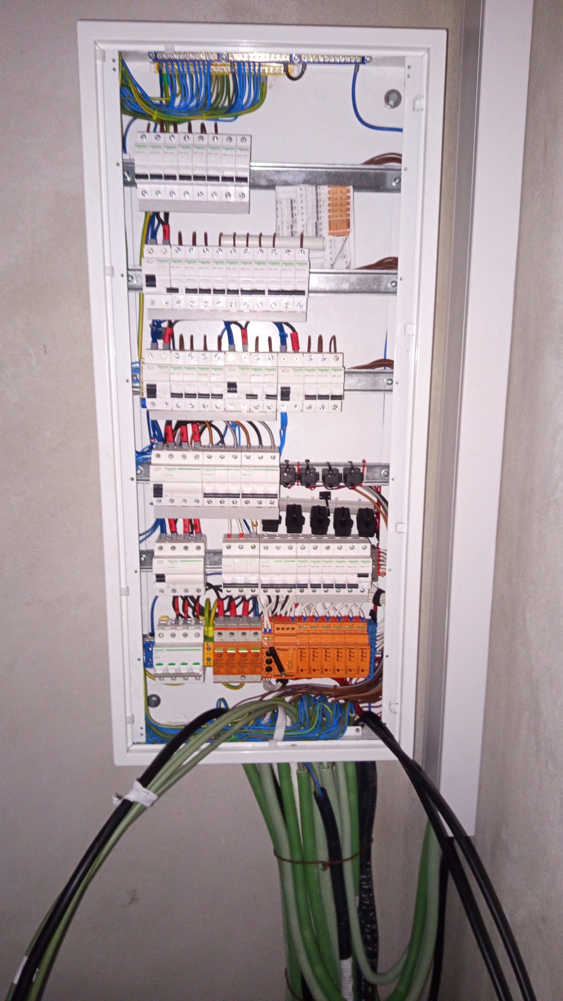 Rozdzielnica elektryczna w białej obudowie, widoczne liczne przewody, bezpieczniki i elementy montażowe. U dołu wiązka kabli w zielonej i czarnej izolacji.