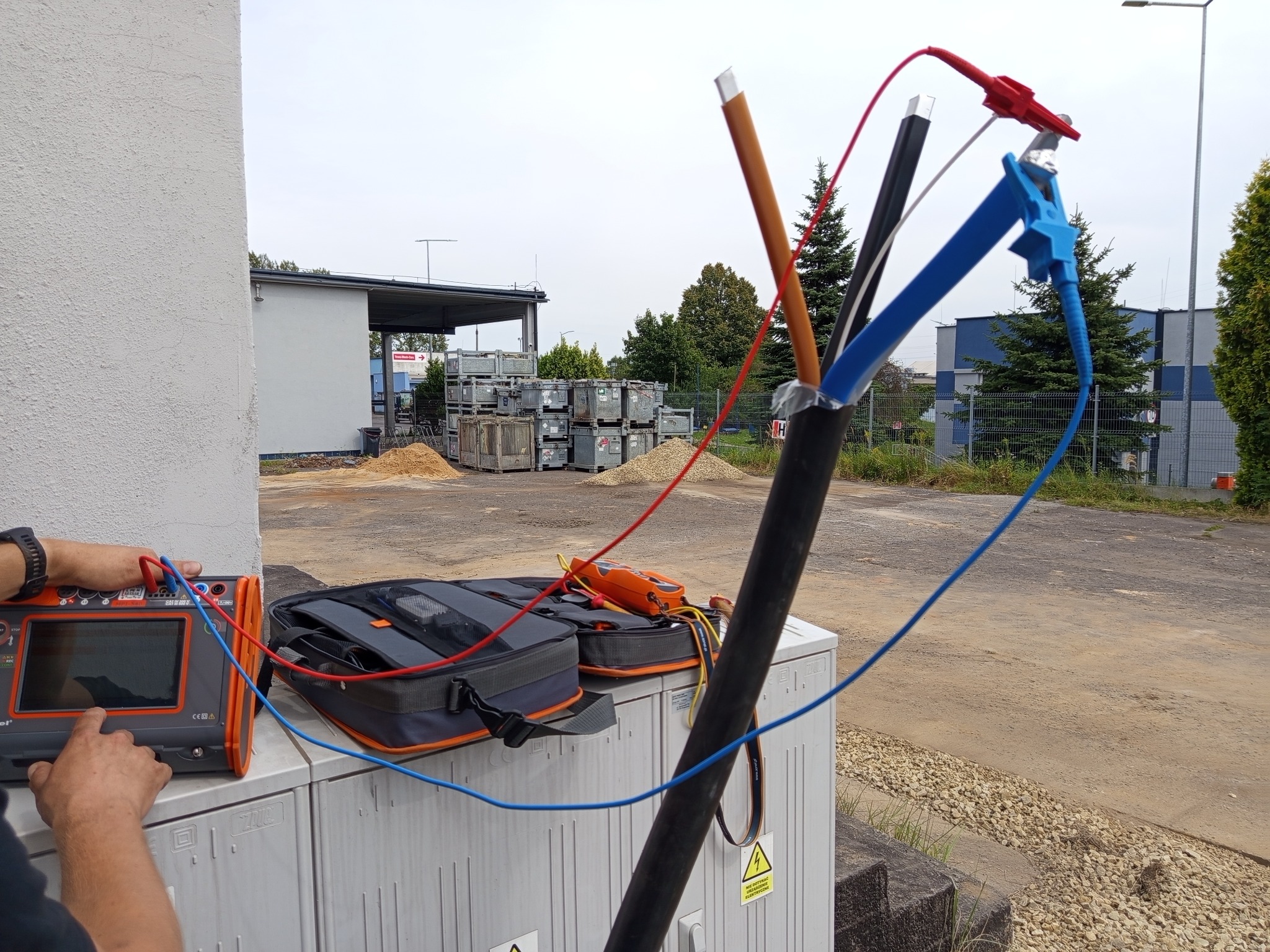 Pomiary elektryczne: kabel z podłączonymi przewodami pomiarowymi i miernikiem na zewnątrz budynku. Widoczne narzędzia i skrzynki narzędziowe.