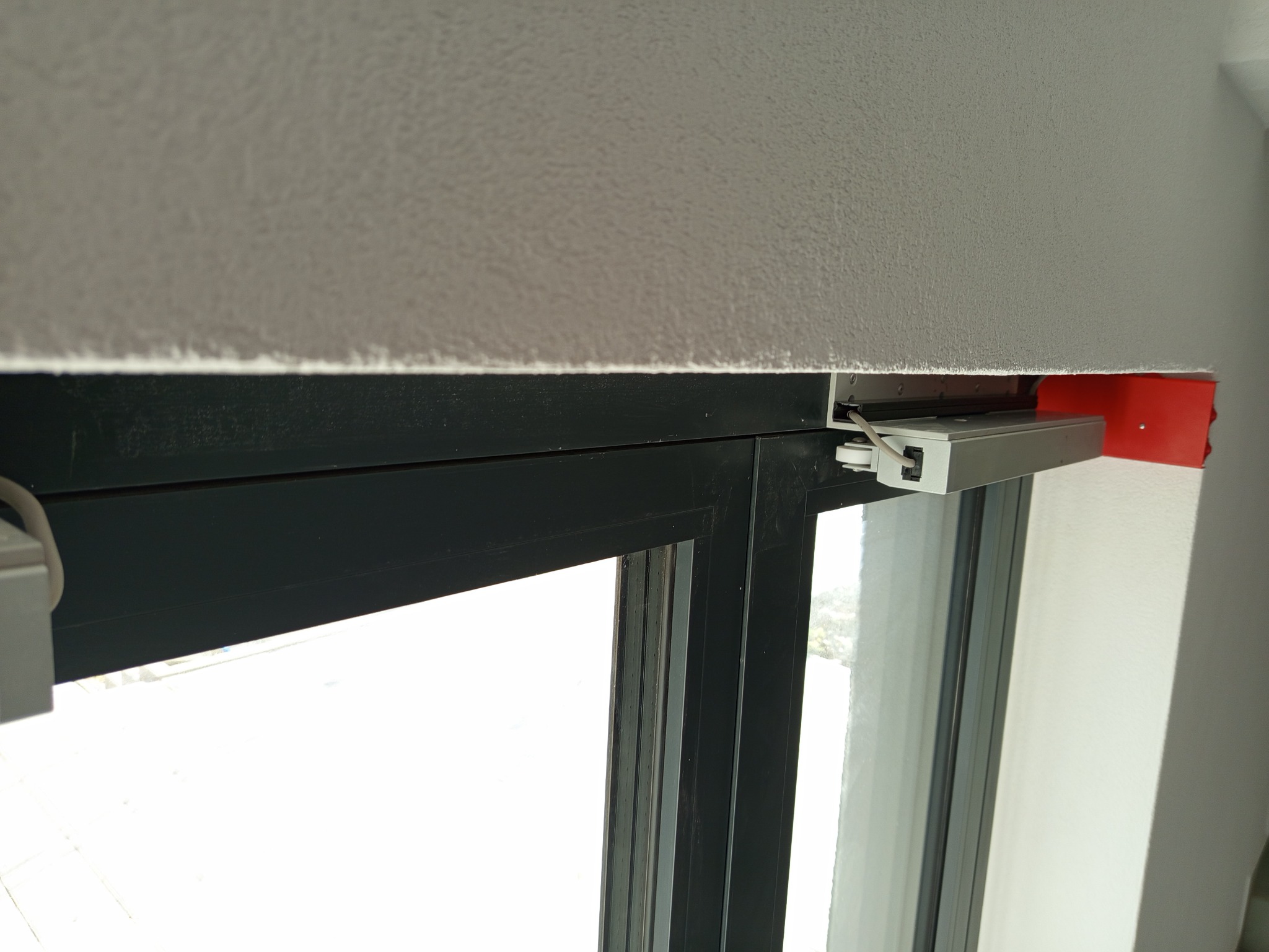 Zamontowany napęd elektryczny okna, widoczny mechanizm i przewody. Ciemna rama okienna kontrastuje z jasnym tłem. Czerwony element obudowy napędu dodaje akcentu kolorystycznego.