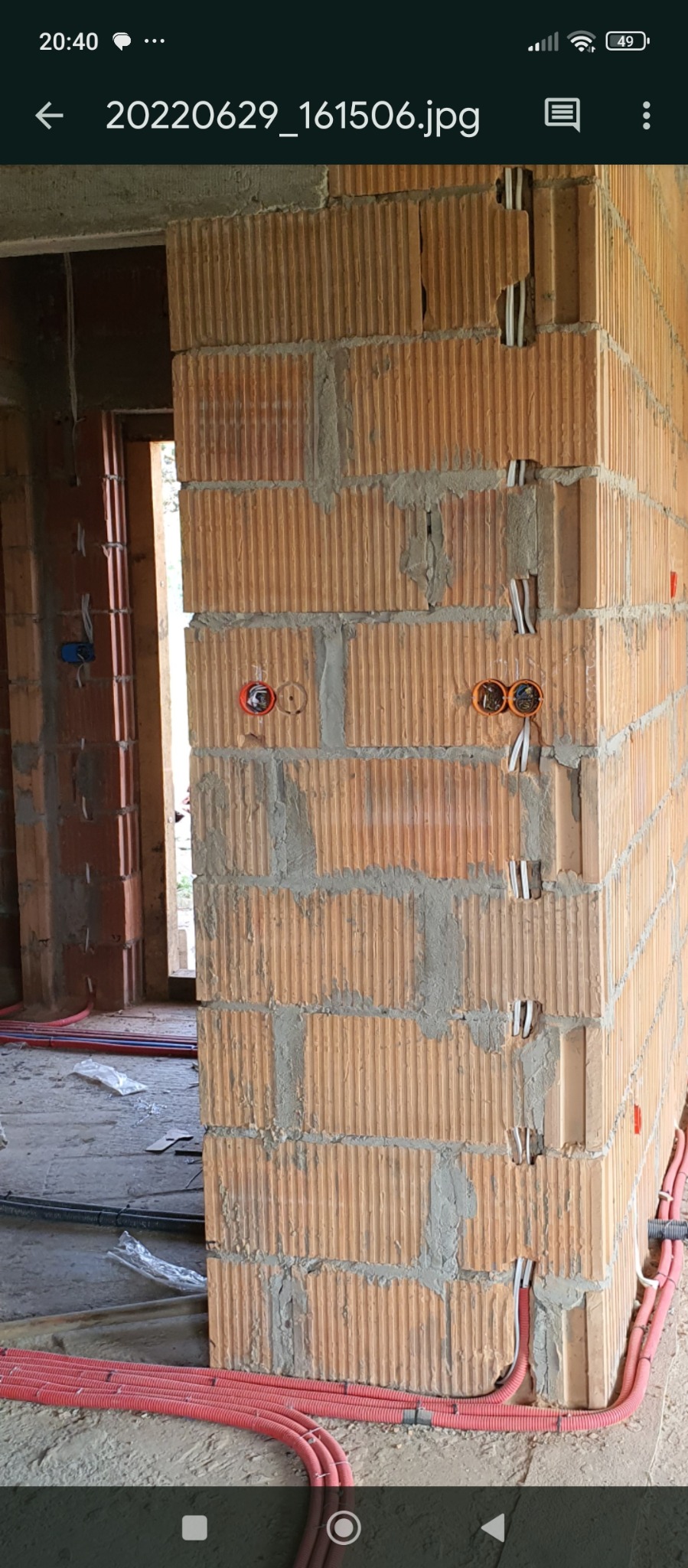 Ściana z pustaków z instalacją elektryczną w trakcie montażu. Widoczne puszki i peszle w kolorze czerwonym. Surowy stan budynku w tle.