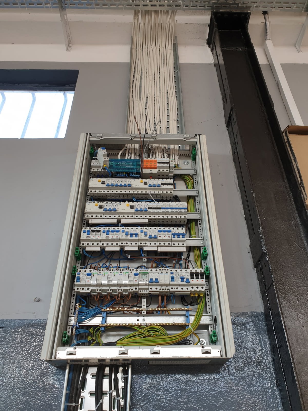 Rozdzielnica elektryczna z licznymi zabezpieczeniami i przewodami, zamontowana na szarej ścianie w pomieszczeniu przemysłowym. Widoczne kable w korytkach.