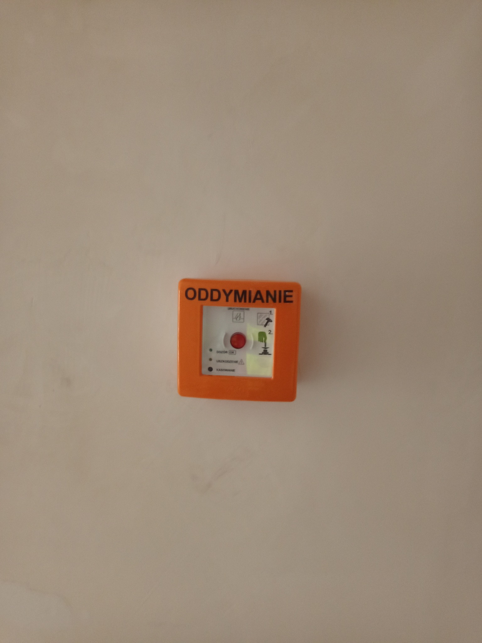 Pomarańczowy przycisk oddymiania na beżowej ścianie. Przycisk z instrukcją obsługi i diodami kontrolnymi. Widok z przodu, centralnie umieszczony.