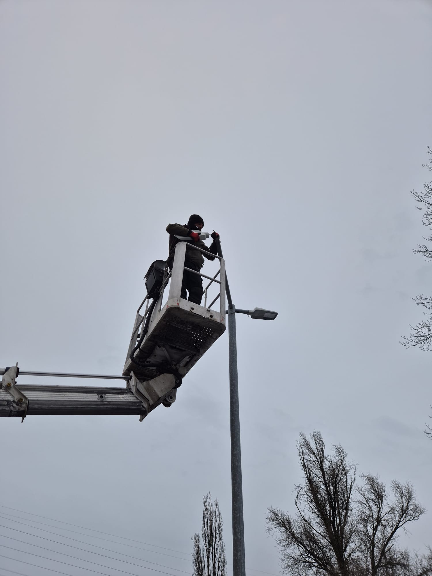 Pracownik w podnośniku koszowym, w masce i rękawicach, montuje lub naprawia oświetlenie uliczne. Szare niebo w tle, widoczne gałęzie drzew.