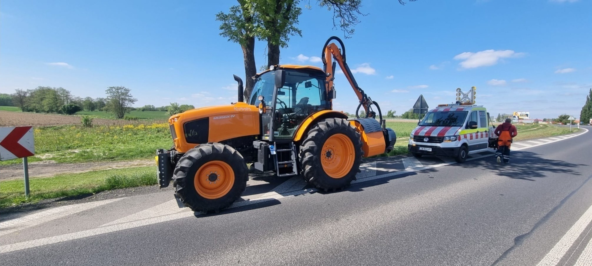 Pomarańczowy traktor Kubota M135GX-IV z zamontowaną głowicą tnącą na poboczu drogi, obok stoi oznakowany bus serwisowy z pracownikiem w kombinezonie.