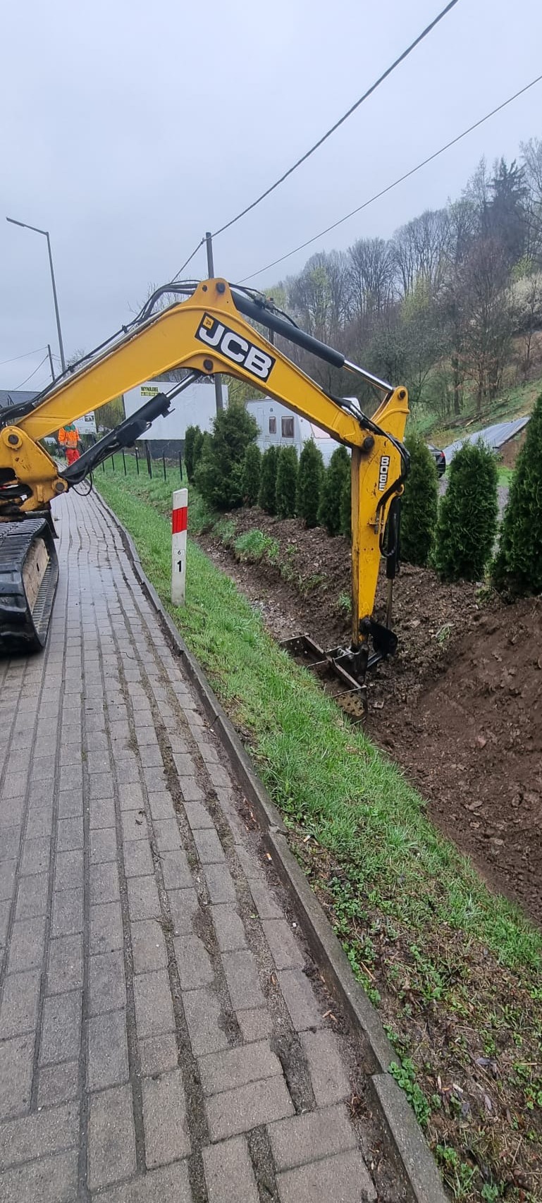 Żółta koparka JCB wykonuje wykop w ziemi obok chodnika z kostki brukowej, w tle rząd tuj i budynki mieszkalne.