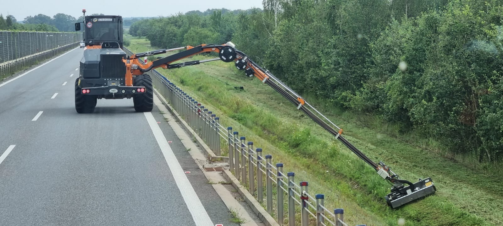 Specjalistyczna maszyna z wysięgnikiem koszącym pobocze drogi ekspresowej, widok zza barierek.