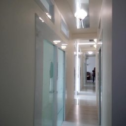 USŁUGI REMONTOWO-BUDOWLANE WIESŁAW BLOCH - Długi, wąski korytarz z białymi ścianami i marmurową podłogą, oświetlony przez szereg lamp wbudowanych w sufit. Po lewej stronie widoczne szklane drzwi z białymi ramami. W głębi korytarza widoczna...