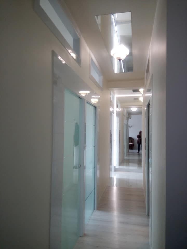 Długi, wąski korytarz z białymi ścianami i marmurową podłogą, oświetlony przez szereg lamp wbudowanych w sufit. Po lewej stronie widoczne szklane drzwi z białymi ramami. W głębi korytarza widoczna...