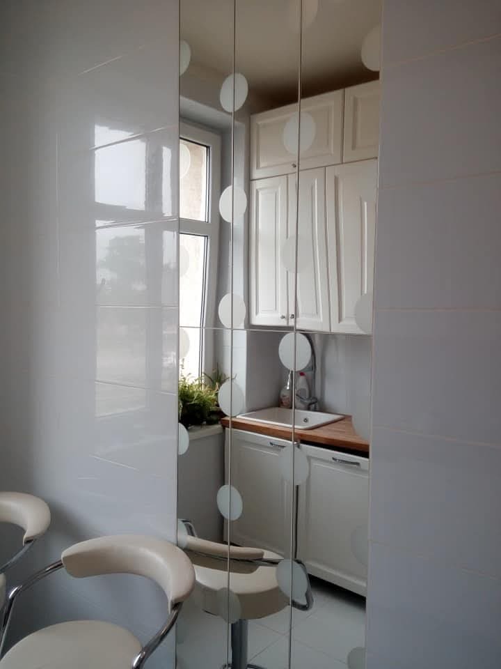 Wykończona, jasna kuchnia z białymi płytkami na ścianach, białymi meblami i drewnianym blatem, widoczna w lustrzanym odbiciu z dekoracyjnymi, okrągłymi naklejkami.