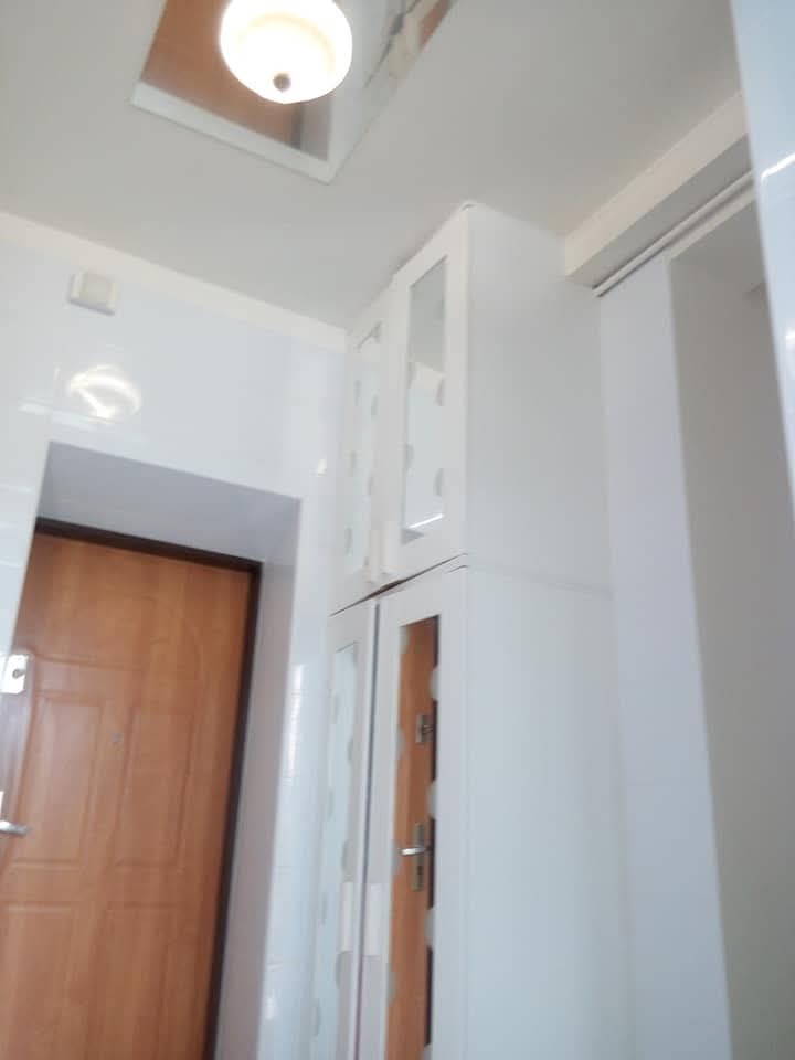 Biała szafka z przeszklonymi drzwiczkami, zamontowana w przedpokoju z brązowymi drzwiami wejściowymi i białymi płytkami ściennymi, oświetlona lampą sufitową.