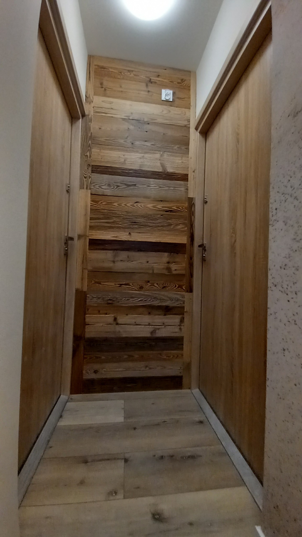 Wąski korytarz z drewnianą ścianą i dwiema drewnianymi drzwiami po bokach, podłoga wyłożona panelami imitującymi drewno, sufit z oświetleniem punktowym.