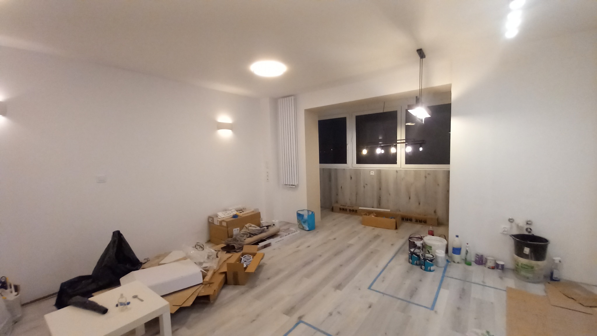 Wnętrze pokoju w trakcie prac wykończeniowych: widoczne białe ściany, podłoga wyłożona jasnymi panelami, okno z widokiem na zewnątrz, kartony, farby i narzędzia malarskie, oraz lampa sufitowa.