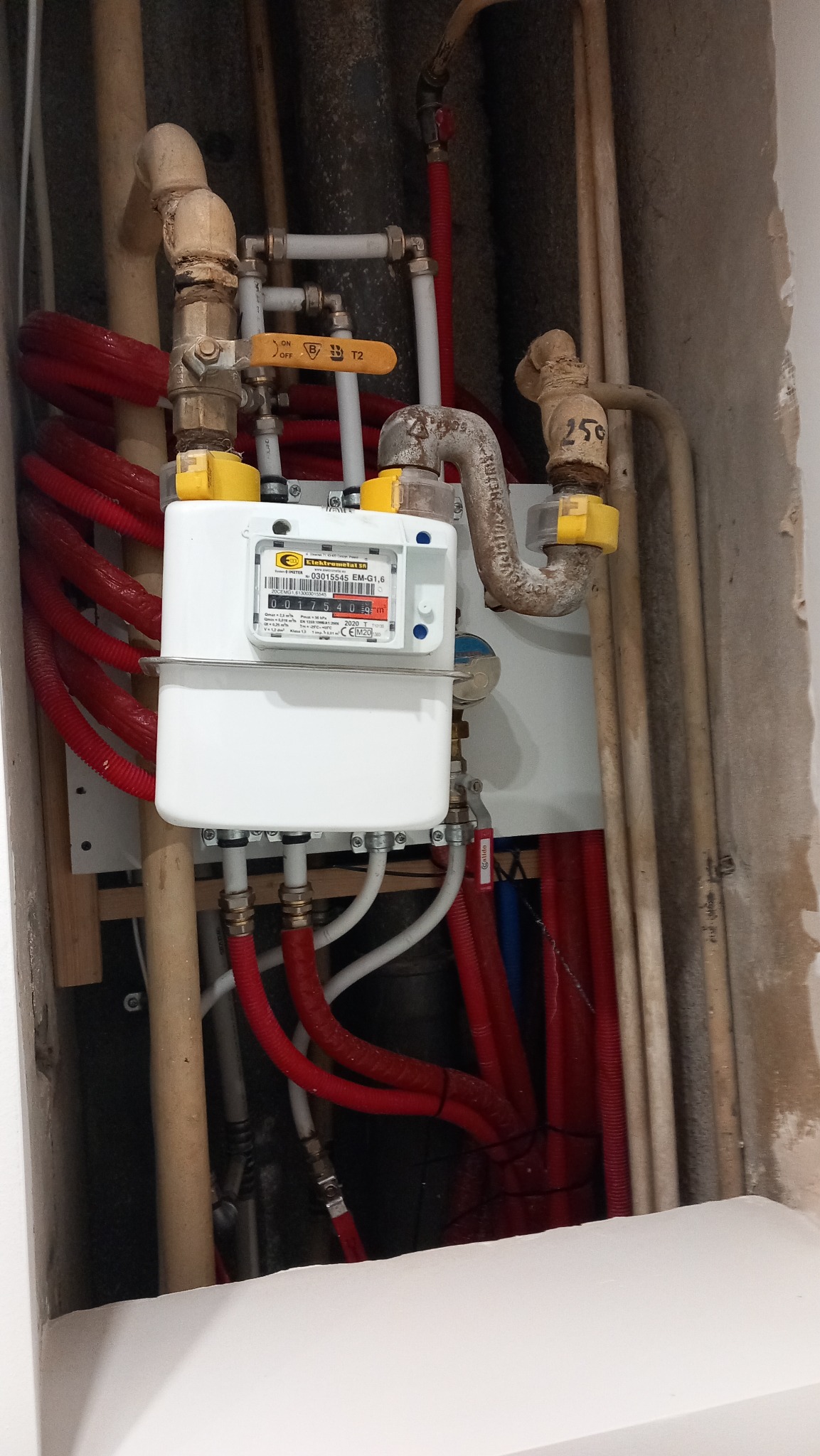 Biały licznik gazu z żółtymi zaworami zamontowany na ścianie, widoczne rury miedziane i plastikowe w otworze w ścianie, szare ściany w stanie surowym.